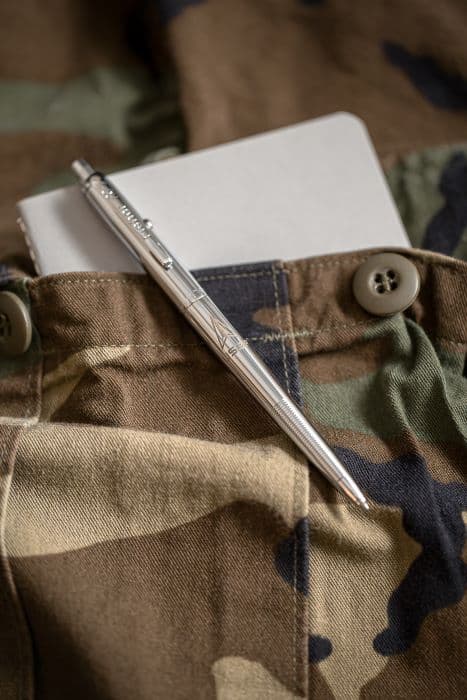 Chrome AG7-SFD Original Astronaut Space Pen with US Space Force insignia laying atop a camouflage pocket with a notebook inside it.