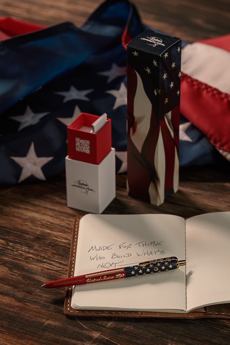 American flag-themed AG7 Space Pen from the America 250 Collection displayed on an open notebook beside patriotic packaging and a folded U.S. flag, with handwritten text reading “Made for those who build what’s next.”