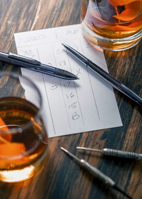 Open 400BTN black titanium nitride bullet space pen on the right. To the left is an open black titanium nitride Infinium space pen. Beneath the pens is a score sheet. Next to the pens are 2 darts and 2 glasses holding an amber colored drink.