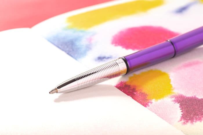 Open 400PP purple bullet space pen laying atop a notebook with printed red, pink, yellow, and blue watercolors