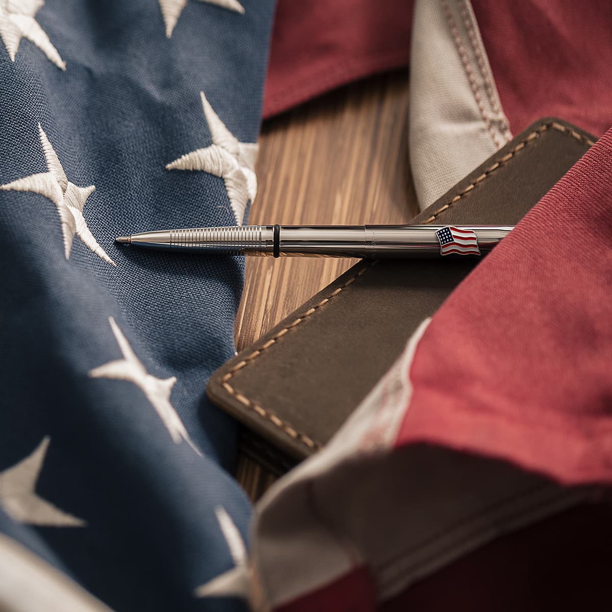 Open chrome x-mark bullet space pen with american flag emblem on cap laying atop a brown notebook and a US flag above a wood surface