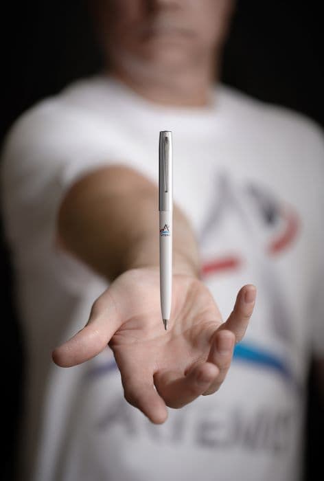 White cap-o-matic space pen with chrome cap, clip, and tip imprinted with Artemis logo appearing to hover over an outstretched hand.