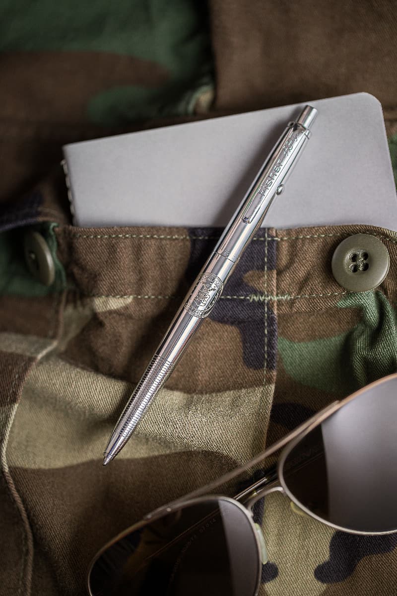 AG7-AF Original Astronaut Space Pen in chrome laying atop a camouflage pocket. There is a notebook in the pocket and a pair of aviator sunglasses below the pen.
